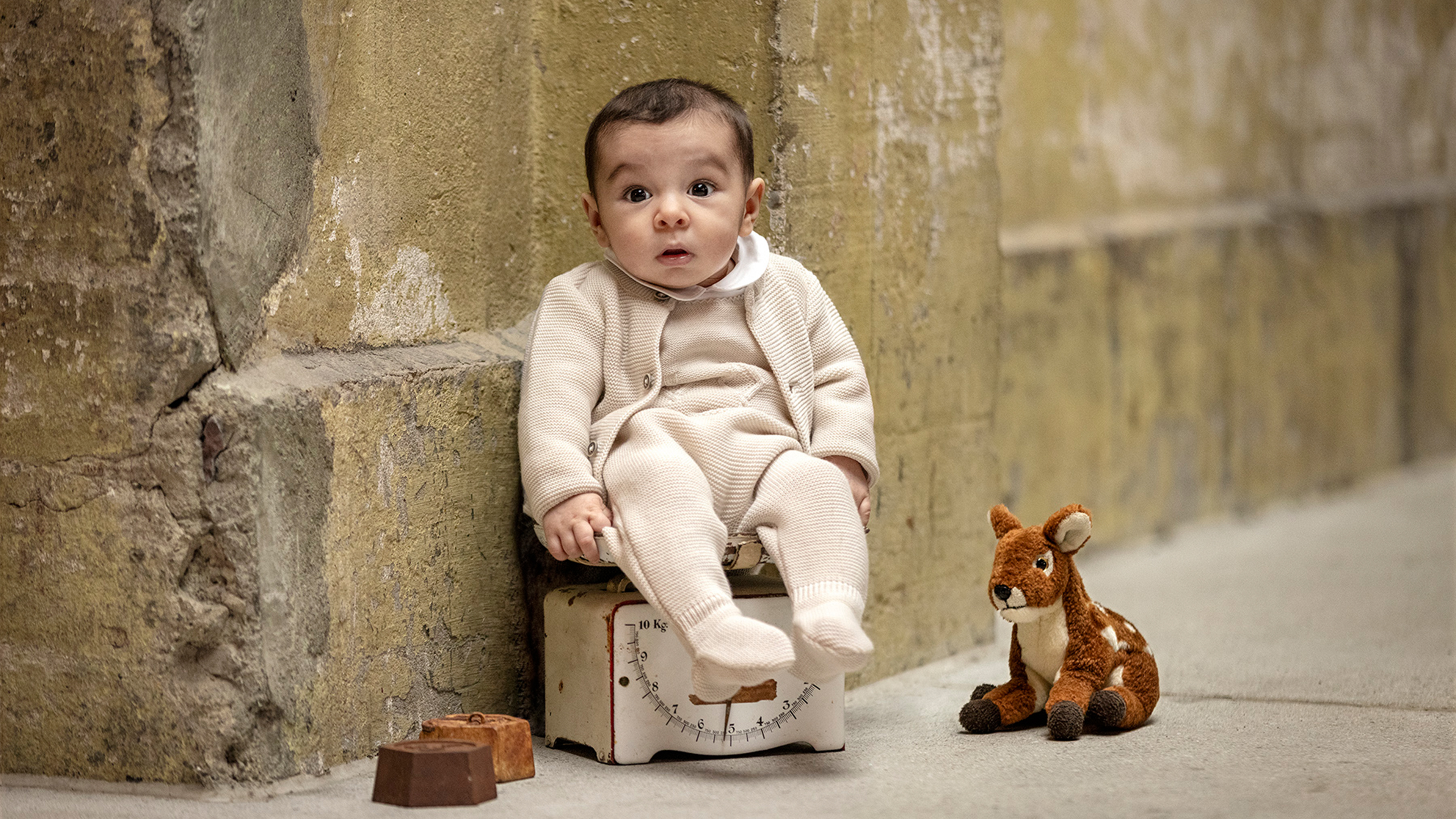 Baby in beiger Strickkleidung auf einer Spielzeugwaage sitzend