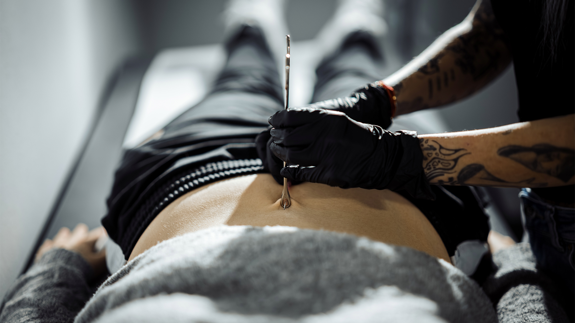 Professioneller Piercer setzt mit einer Piercingzange ein Bauchnabelpiercing bei einer Kundin in einem professionellen Piercingstudio.