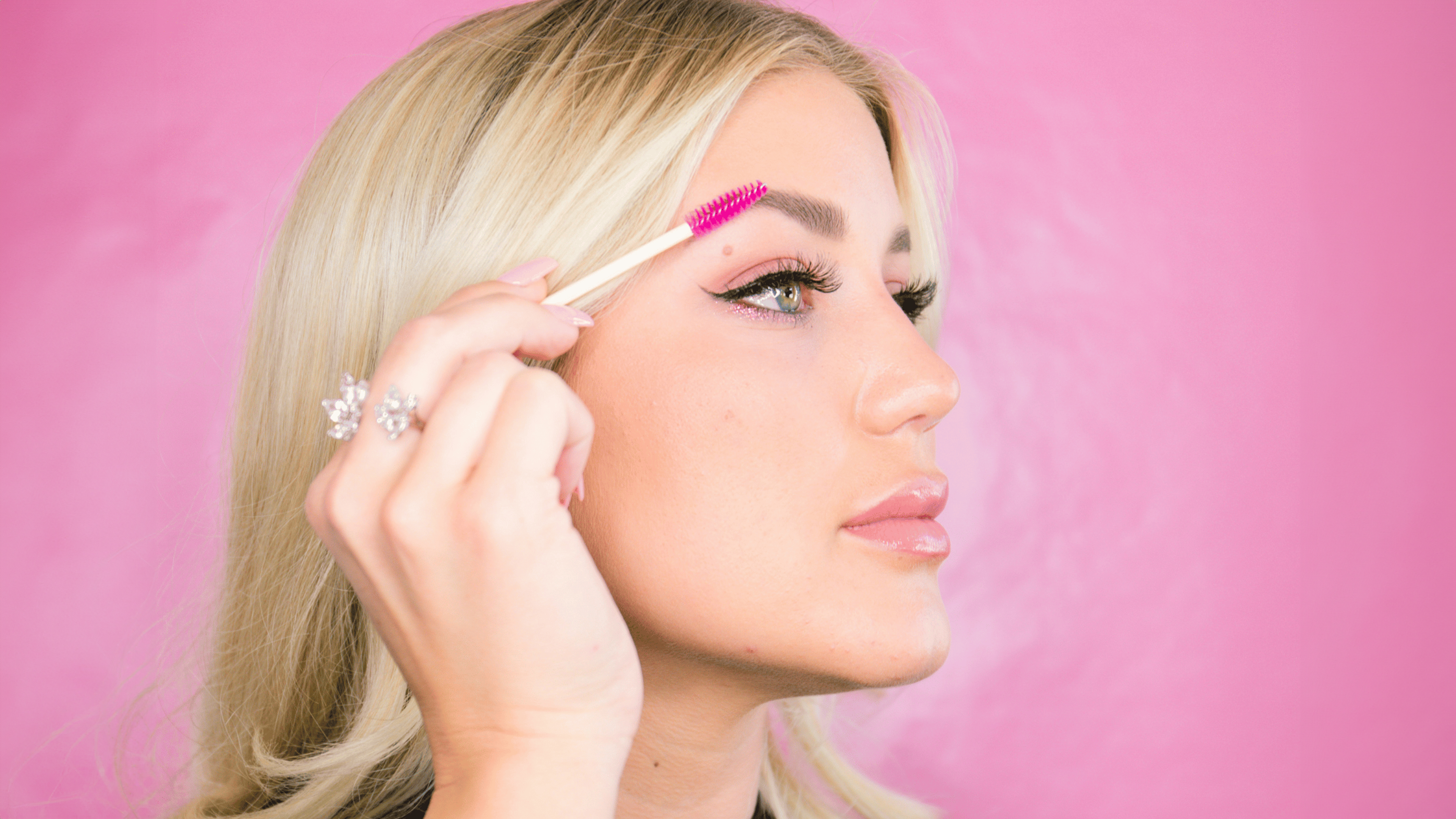 Blonde Frau mit pinkem Wimpernbürstchen vor pinker Wand