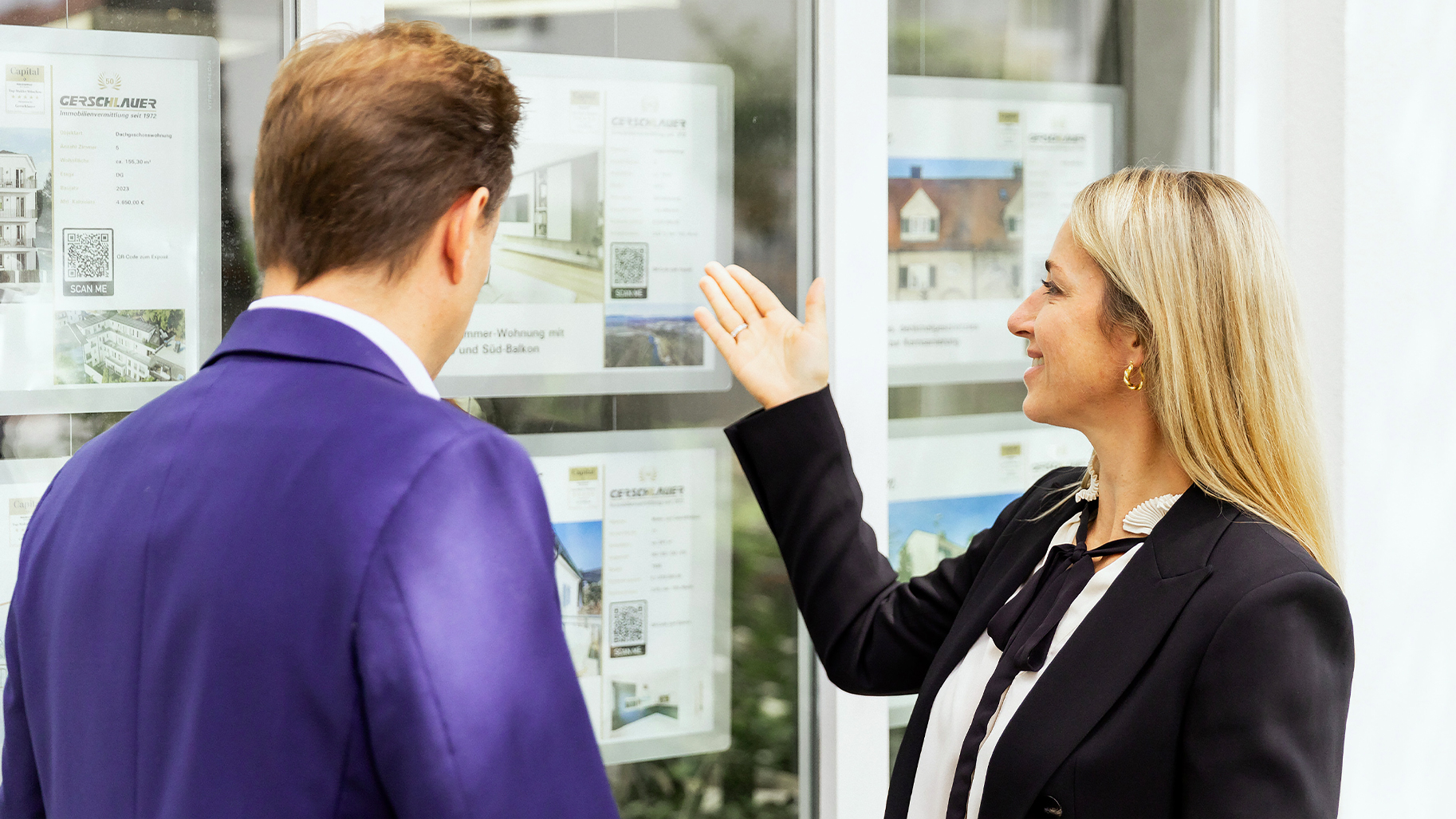 Eine Immobilienmaklerin von Gerschlauer Immobilien in München zeigt einem Kunden aktuelle Immobilienangebote im Schaukasten.