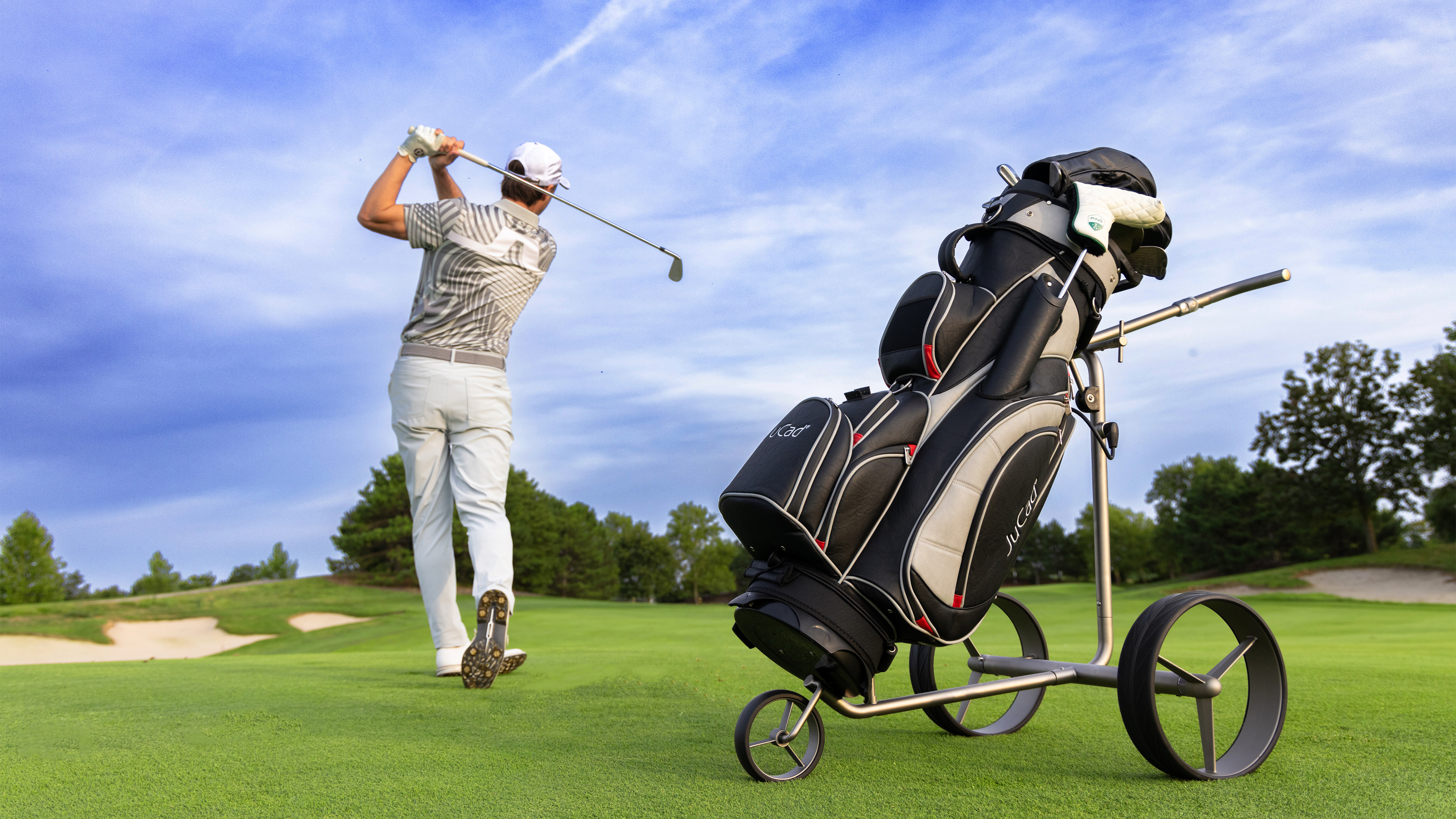 Mann im Hintergrund spielt Golf, im Vordergrund ein Golftrolley