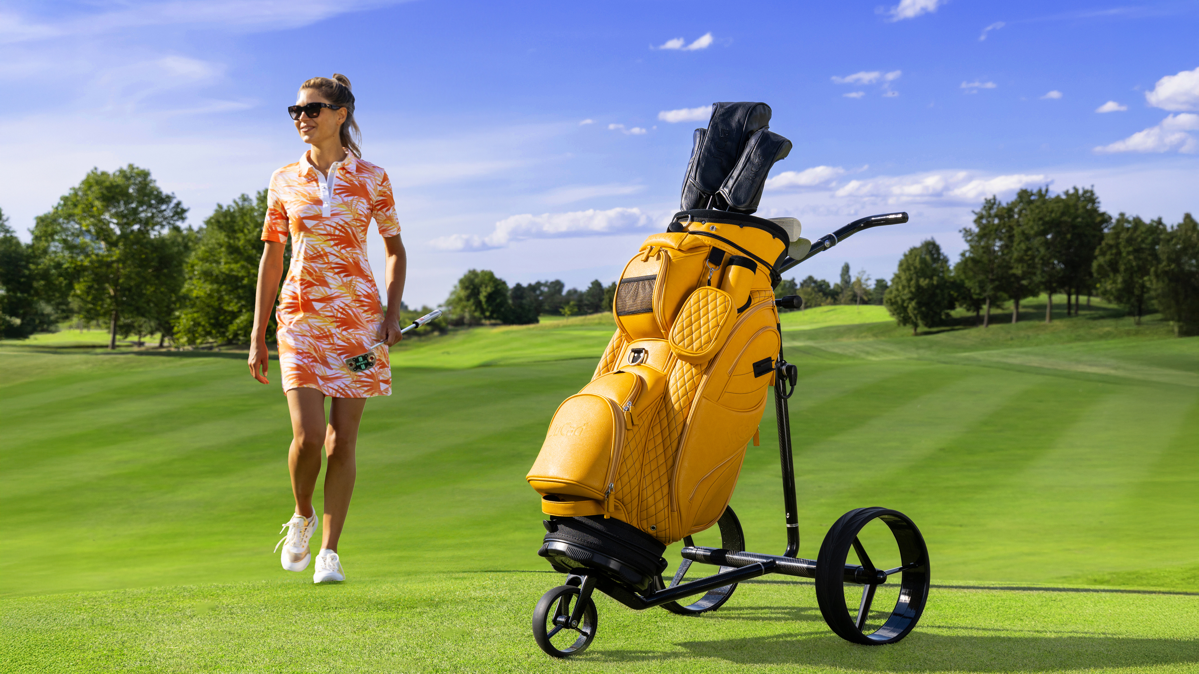 Frau im Hintergrund auf einem Golfplatz, im Vordergrund gelber Golftrolley