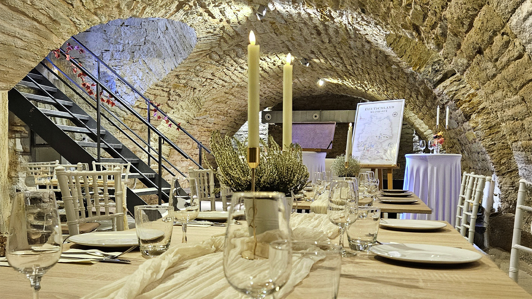 Perspektivische Ansicht einer festlich gedeckten Tafel im historischen Gew&ouml;lbekeller von genusshochzwei. Das warme Kerzenlicht und die rustikalen Natursteinb&ouml;gen schaffen eine intime Atmosph&auml;re f&uuml;r exklusive Wein-Events und Experten-Verkostungen mit Sommelier-Begleitung.