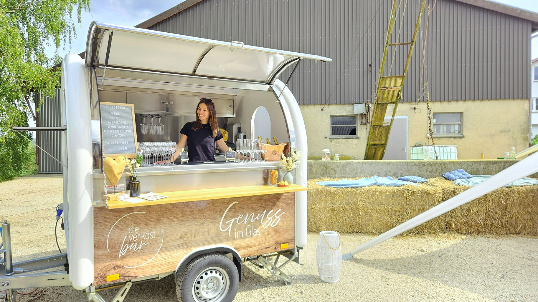 Die mobile VerkostBAR von genusshochzwei bei einem Outdoor-Event. Eine l&auml;chelnde Eventmanagerin ber&auml;t G&auml;ste an dem stilvollen, wei&szlig;en Bar-Anh&auml;nger mit der Aufschrift &sbquo;Genuss im Glas&lsquo;. Im Hintergrund schaffen Heuballen und eine l&auml;ndliche Kulisse ein exklusives, entspanntes Ambiente f&uuml;r besondere Genuss-Events.