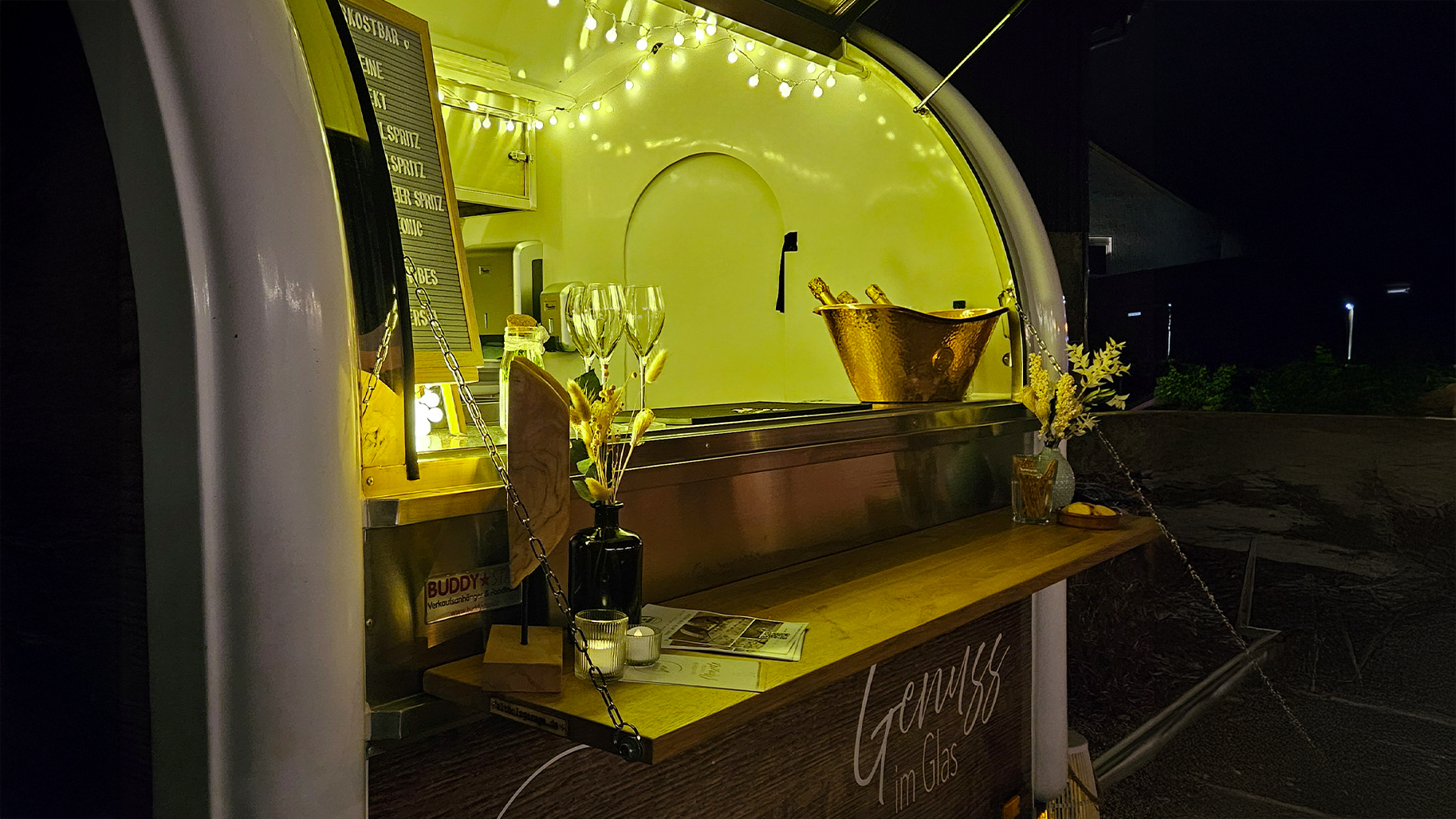 Die beleuchtete VerkostBAR von genusshochzwei bei einem Abend-Event. Der stilvolle Bar-Anh&auml;nger mit Lichterketten, Sektk&uuml;hler und Weingl&auml;sern schafft eine exklusive Atmosph&auml;re f&uuml;r Genuss-Events. Perfekt inszeniertes mobiles Catering f&uuml;r Weinverkostungen und besondere Anl&auml;sse bei Nacht.