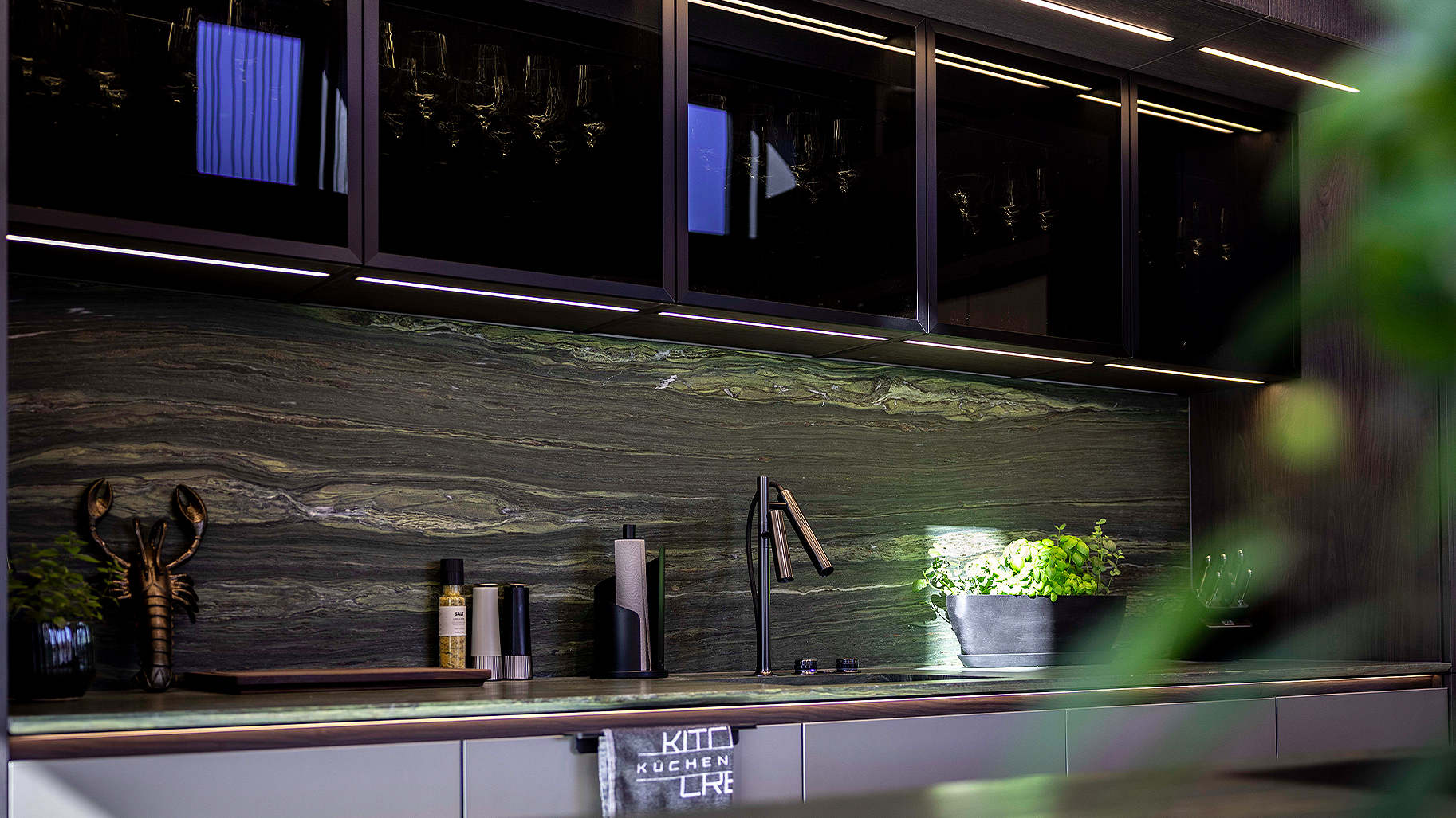 Dunkle Premium-Küche mit Glasfronten, indirekter Beleuchtung und edler Rückwand – Kitchen Crew Küchendesign