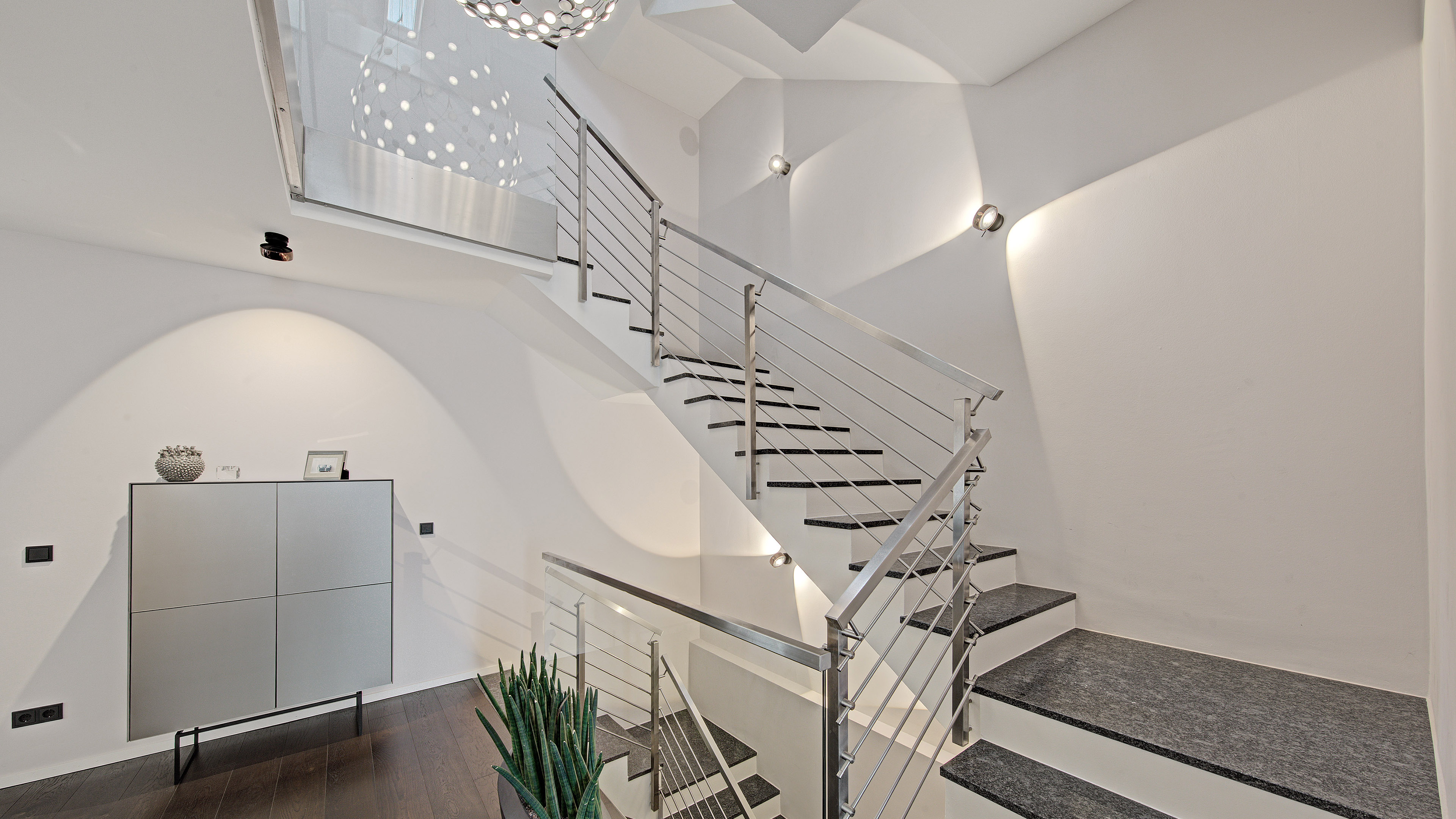 Modernes Treppenhaus mit Edelstahlgeländer, dunklen Stufen und gezielt gesetzten Wand- und Deckenleuchten.