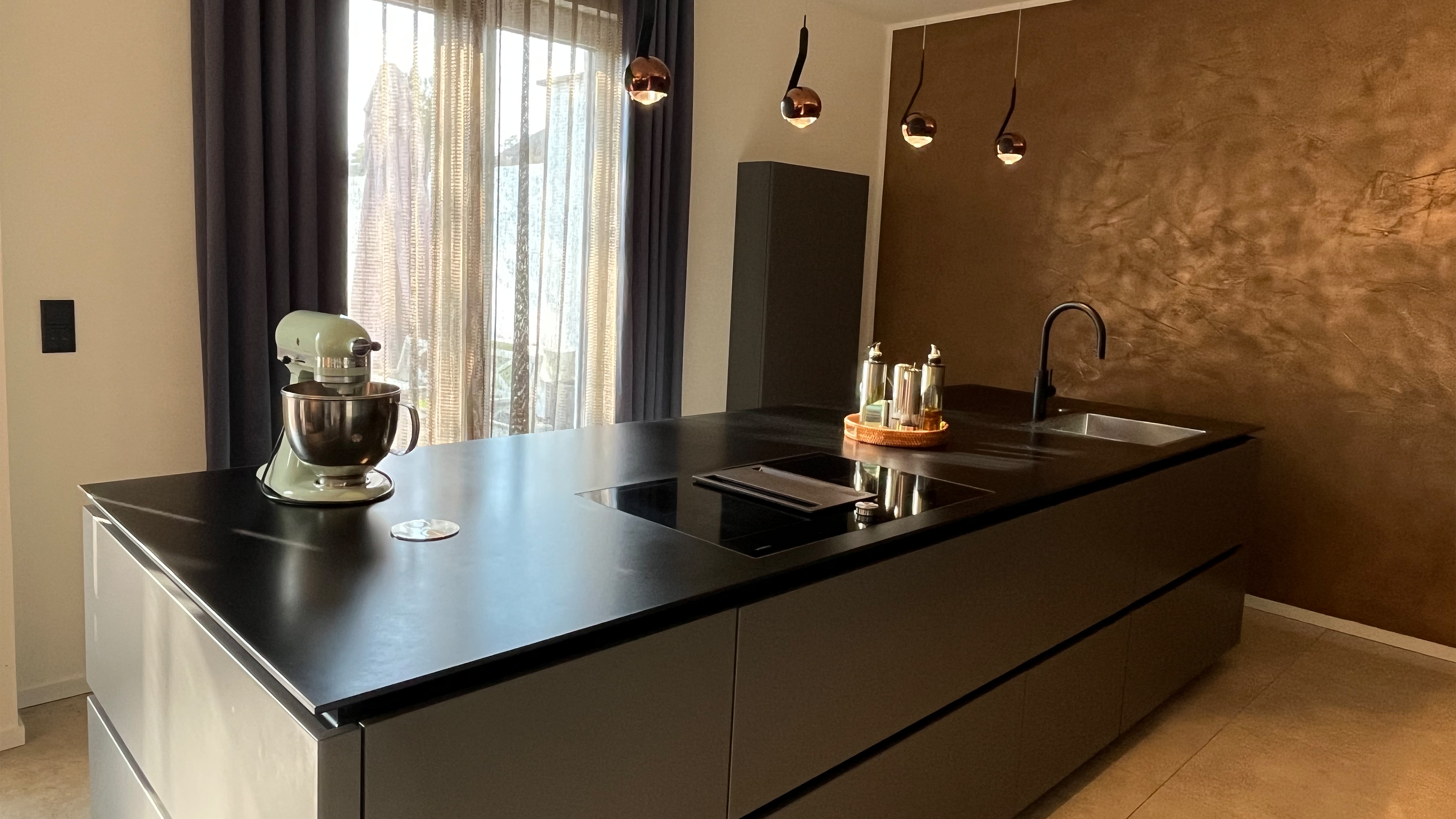 Moderne Küche mit schwarzer Kochinsel, grauen Einbauschränken, Kupferwand und skulpturalen Pendelleuchten.