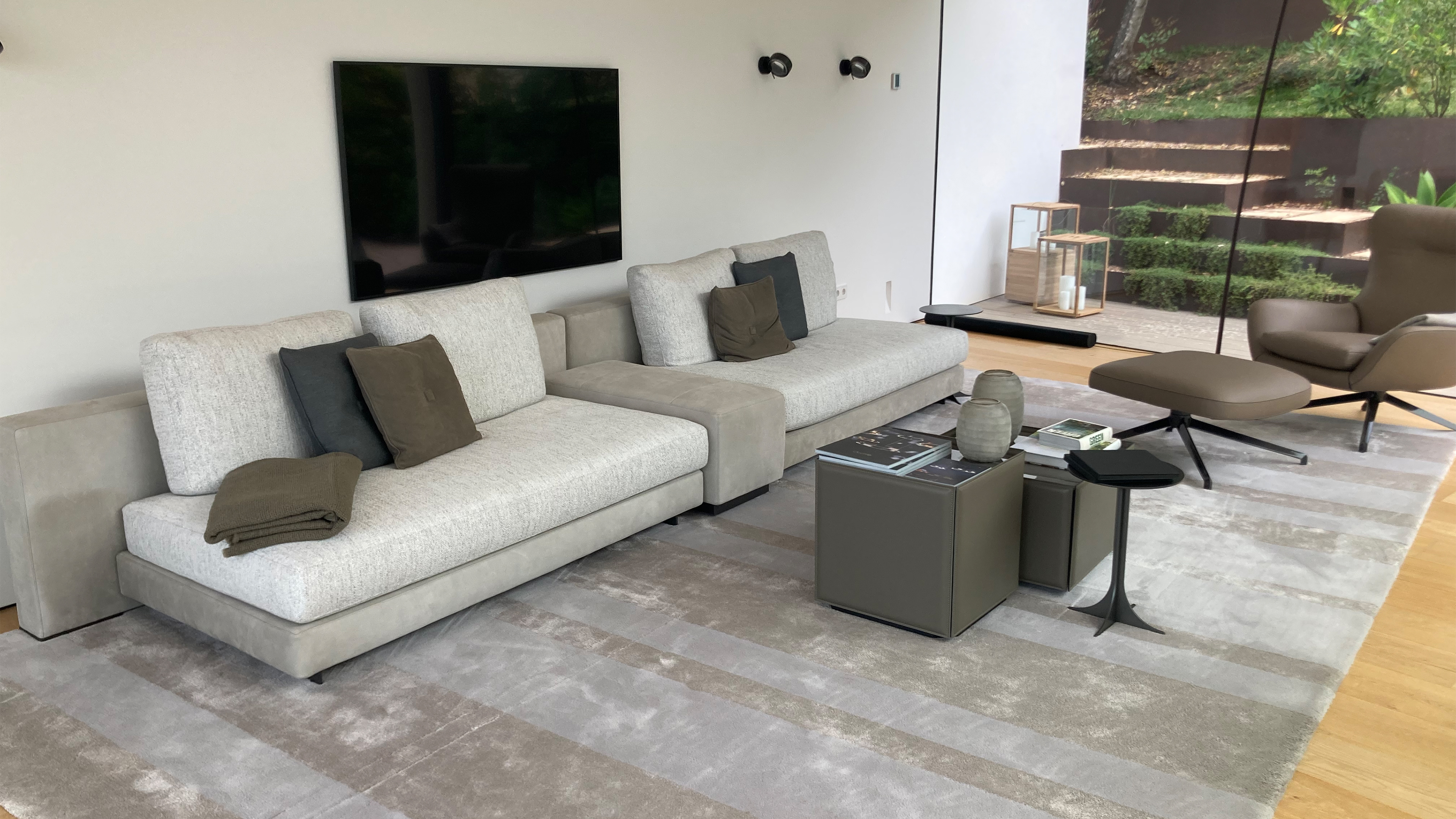 Modernes Wohnzimmer mit modularem Sofa, wandmontiertem Fernseher, Soundbar und raumhoher Verglasung zum Garten.