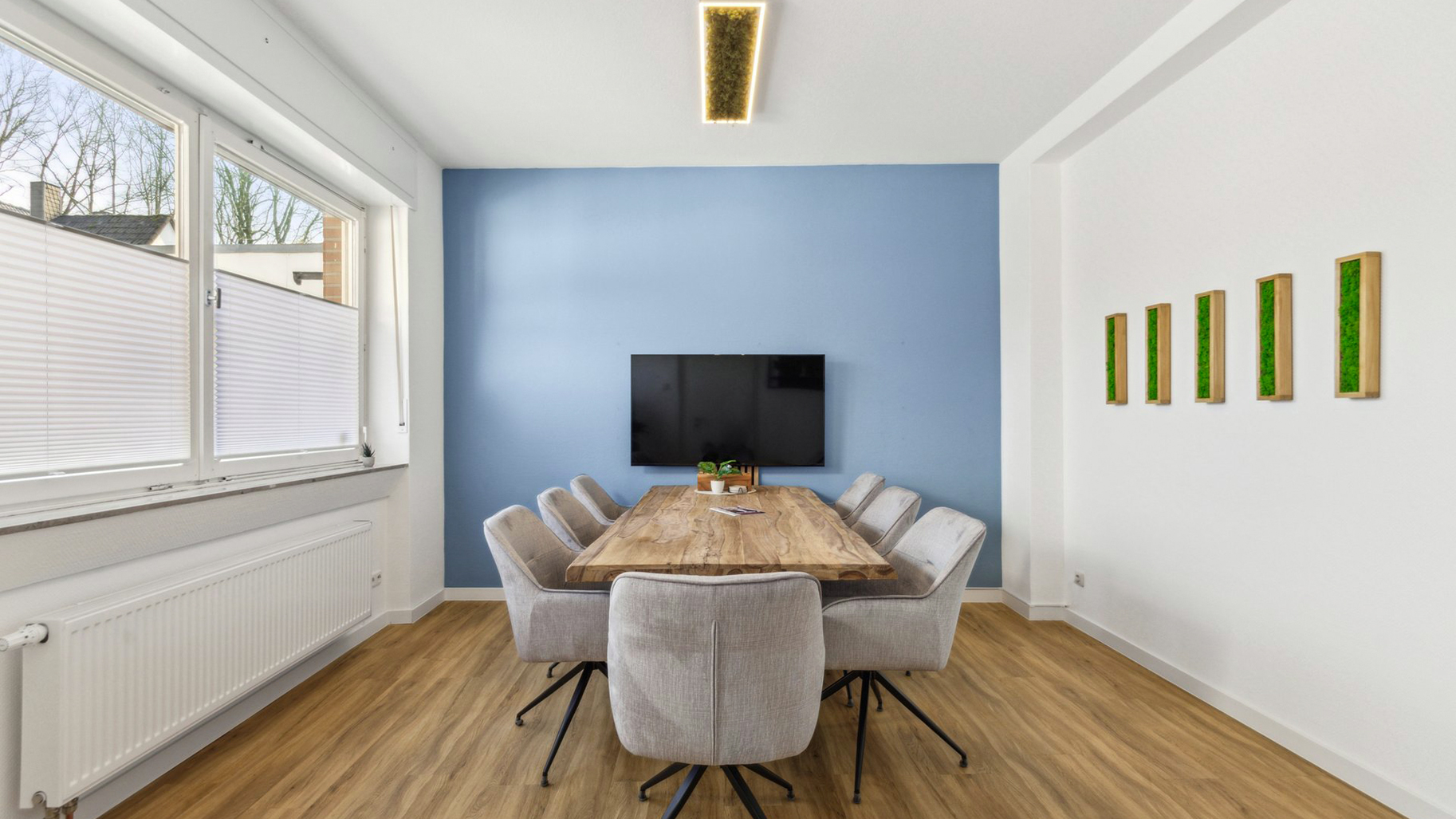 Moderner Besprechungsraum mit Holztisch, grauen Stühlen, Bildschirm und blauer Akzentwand.